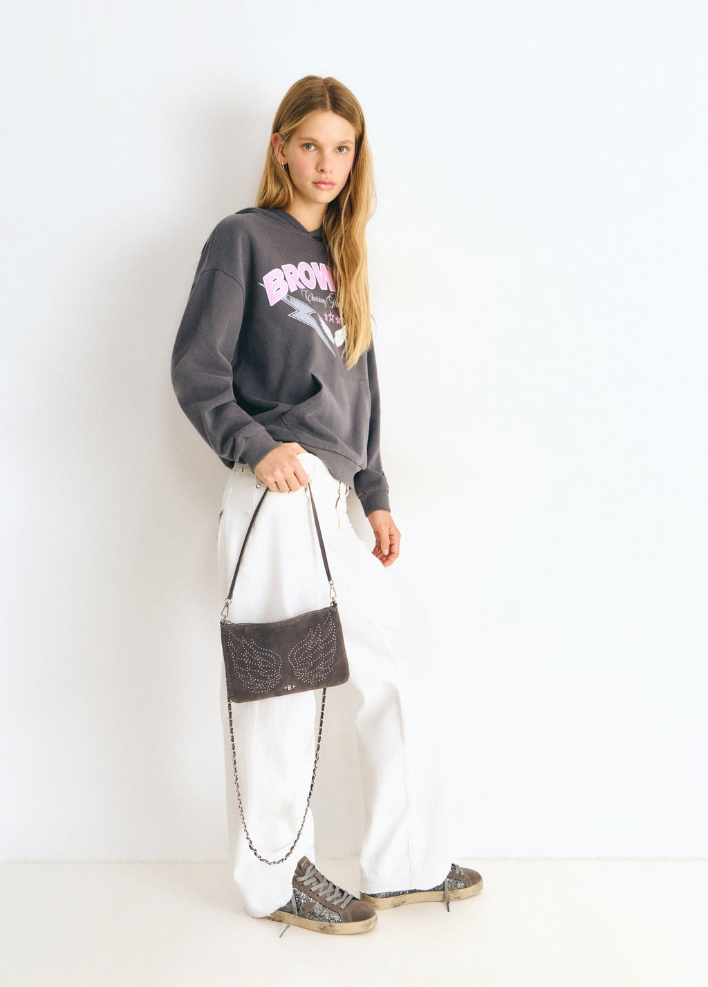 Bolso de color gris con diseño de alas y cadena metálica colgado sobre vaqueros blancos
